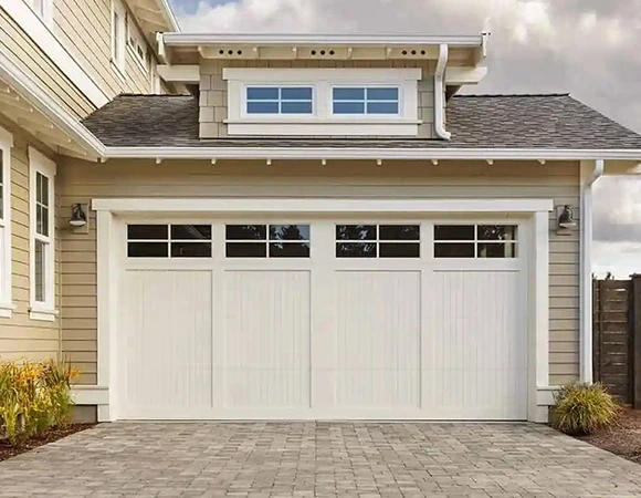 Beige house with a white double garage door, featuring glass windows. The roof has a small dormer window. The foreground shows a stone driveway.