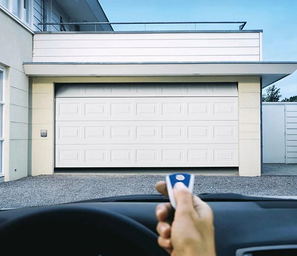 A modern garage door is partially open, viewed from inside a car. A hand holds a remote control, implying automatic operation. The scene is calm and residential.