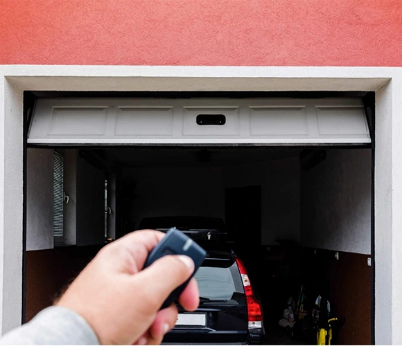 A hand holds a remote control, opening a garage door to reveal a parked car inside. The garage exterior is painted red. The scene feels modern and functional.