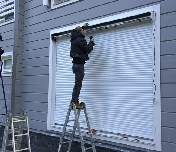 A person in winter clothing stands on a ladder, installing white roller shutters on a gray house. The scene conveys a sense of diligence and focus.
