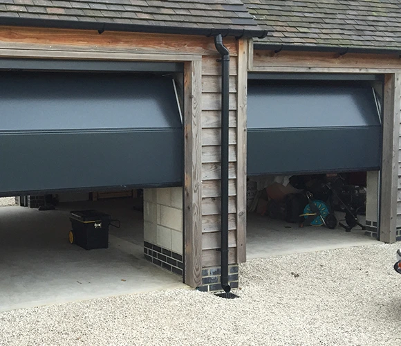 Two adjacent garages with partially open modern grey doors, revealing minimal storage inside. The exterior features wood paneling and gravel.