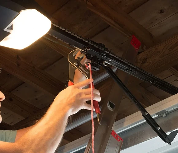 A person uses pliers to repair a ceiling-mounted garage door opener mechanism. The setting is well-lit, with a focused, attentive atmosphere.
