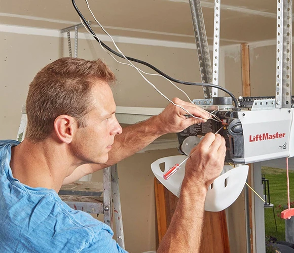 A man in a blue shirt works intently on a LiftMaster garage door opener, using a screwdriver. The setting is a garage, reflecting focus and technical skill.