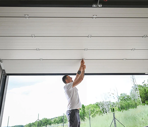 A man in a white shirt stands in a garage, adjusting the overhead door mechanism. He is focused, with greenery visible outside, suggesting a calm setting.