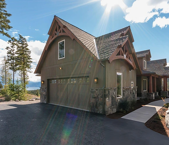 Charming rustic house with gabled roof, stone accents, and large garage. Sunlight streams over a scenic mountain view, creating a warm, serene vibe.