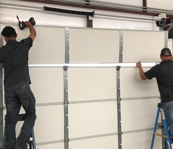 Two men install a white garage door using drills. They stand on ladders, focused on attaching the panels. The setting is a workshop, conveying teamwork.