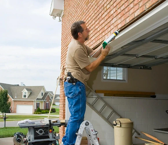 A man standing on a ladder applies sealant to a garage roof. He's focused and working near a brick house in a suburban neighborhood, with tools nearby.