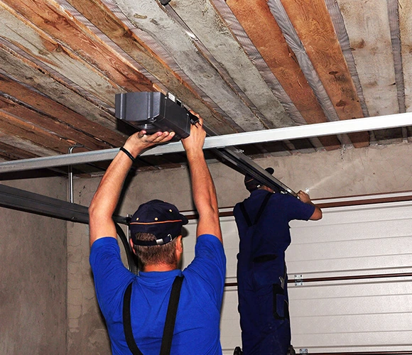 Two workers in blue uniforms install a garage door opener on a wooden ceiling. Their focus suggests concentration and teamwork in a workshop setting.