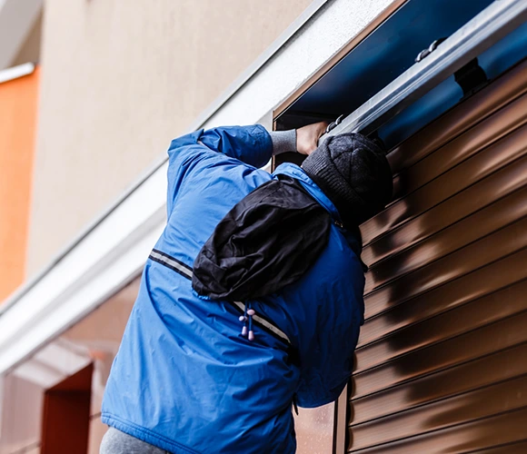 A worker in a blue jacket and black beanie is installing or repairing a brown metal roller shutter on a building exterior, focused and industrious.