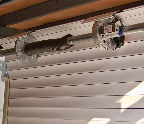 Close-up of a garage door mechanism showing a coiled torsion spring and metal rod. The setting is industrial, with a focus on mechanical function.