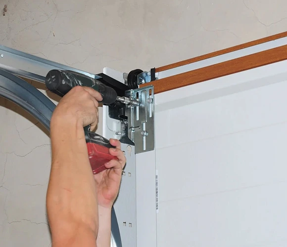 Hands using a power drill to install a metal bracket on a garage door mechanism. The setting is a workshop, conveying focus and precision.