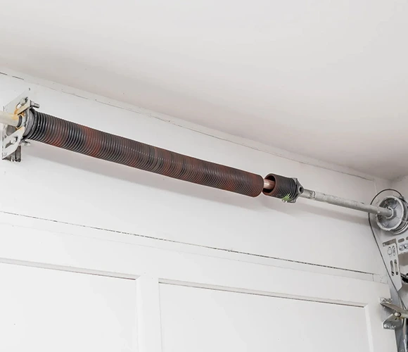 Close-up of a rusty garage door torsion spring mounted on a white ceiling. The metal coil is stretched across the top of the door, showing wear and age.