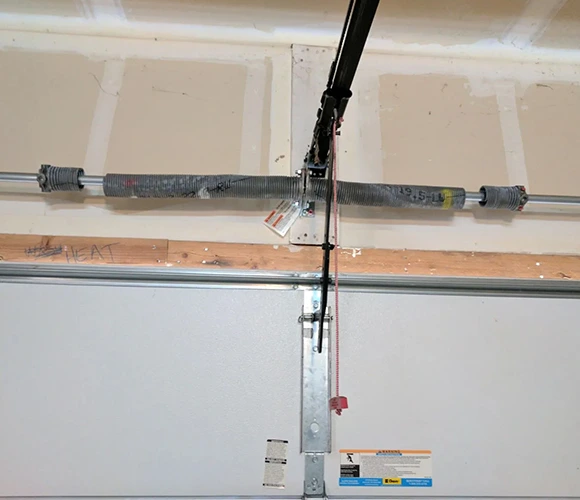 Close-up of a garage door mechanism showing a tension spring on a metal rod. A red cord hangs from the opener. Neutral tones and a utility-focused setting.
