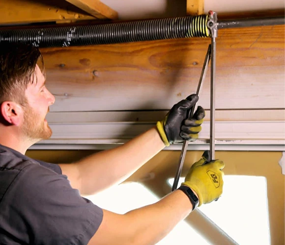 A man wearing work gloves adjusts a garage door torsion spring using tools. He appears focused, working in a well-lit space, conveying a sense of concentration.
