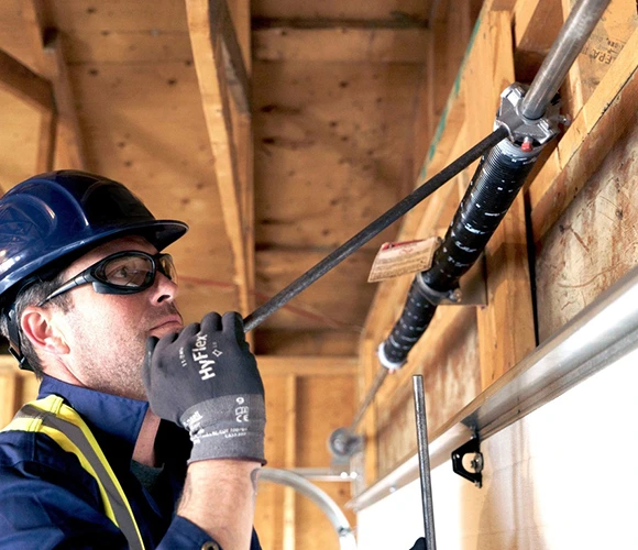 A worker wearing a blue hard hat and safety glasses adjusts a garage door spring. He is focused and uses tools, standing in a wooden-framed structure.