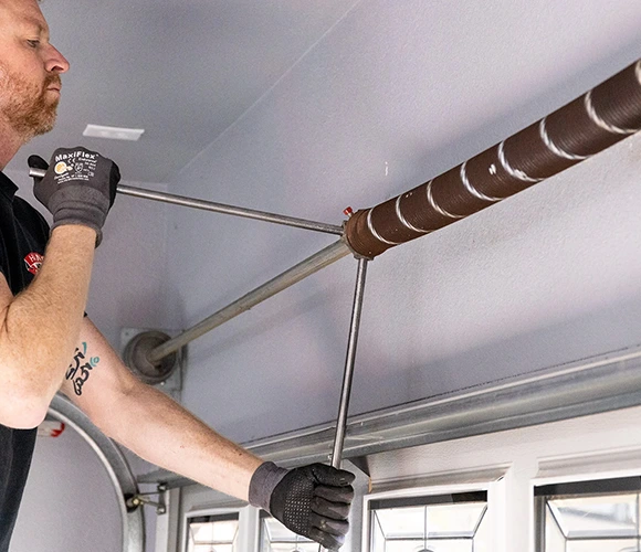 Man adjusting a garage door torsion spring with a steel rod. He wears gloves, focusing intently on the task, conveying a sense of precision.