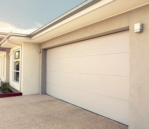 Modern suburban house with a closed beige garage door, light brown exterior, and a few small plants. The scene is bright, with a clear sky.