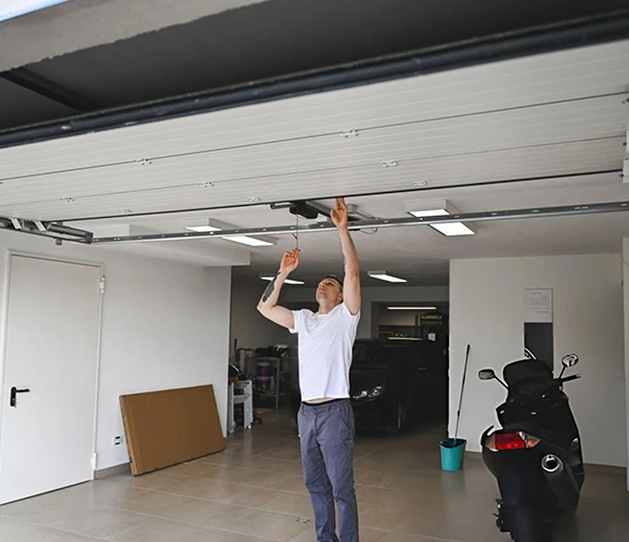 A man in a garage adjusts an overhead door, surrounded by a motorcycle and cleaning supplies. The space is tidy, with a calm, functional atmosphere.